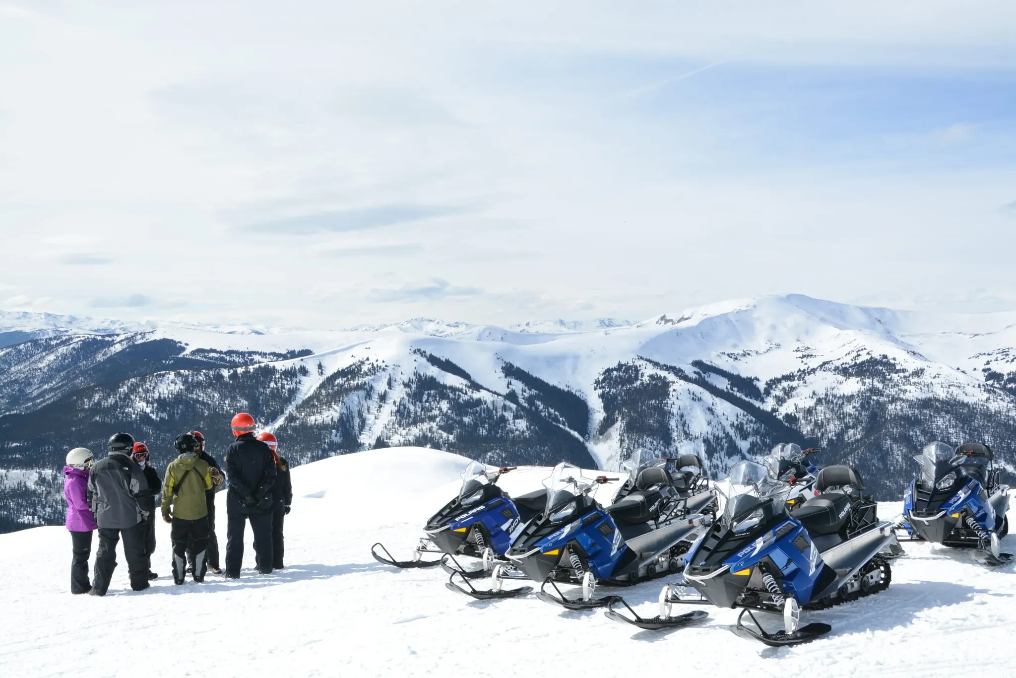 group of riders in a circle standing next to snowmobiles with sweeping mountain views in background.