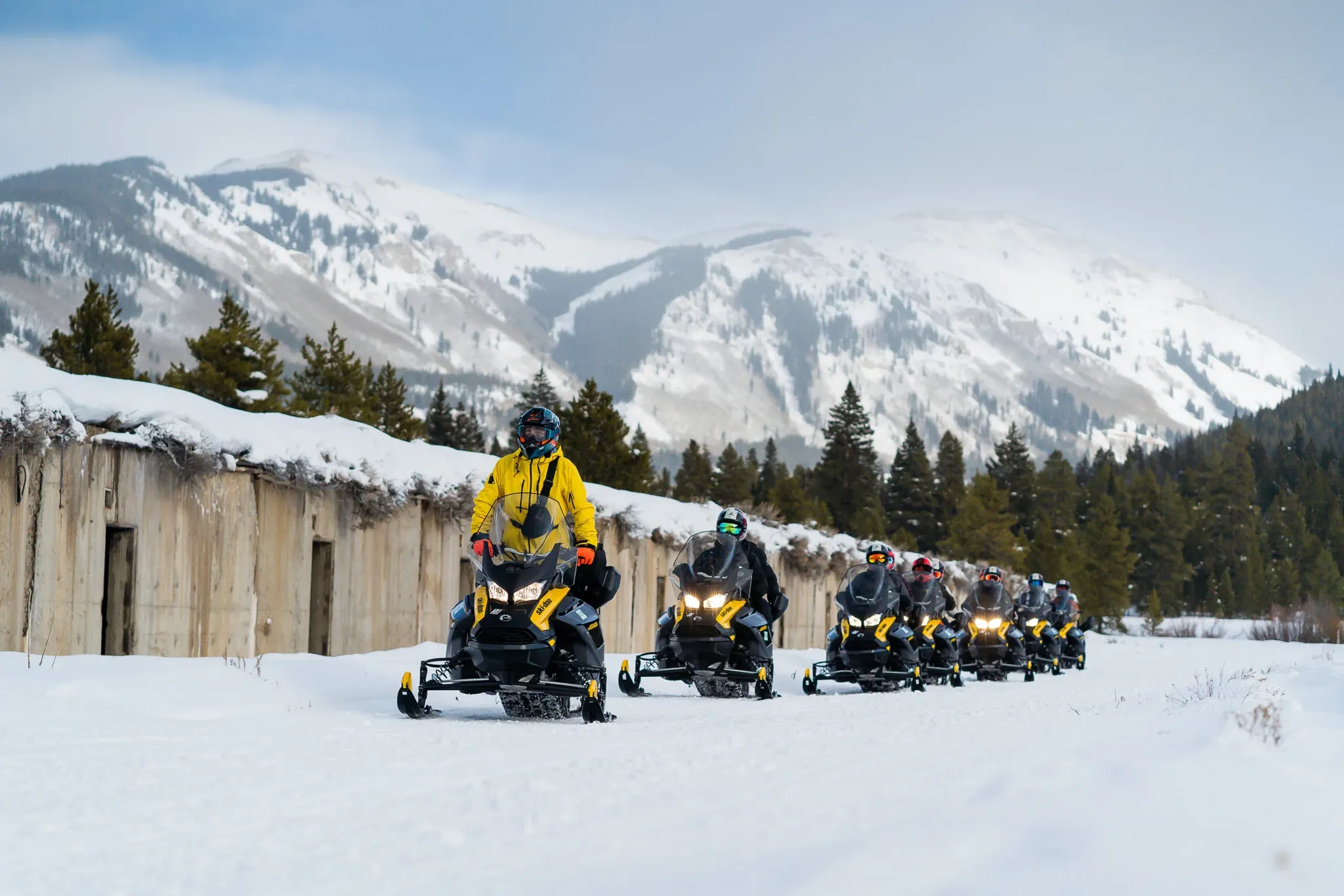 single file line of snowmobilers next to flat top buildings with snowy mountains in background.