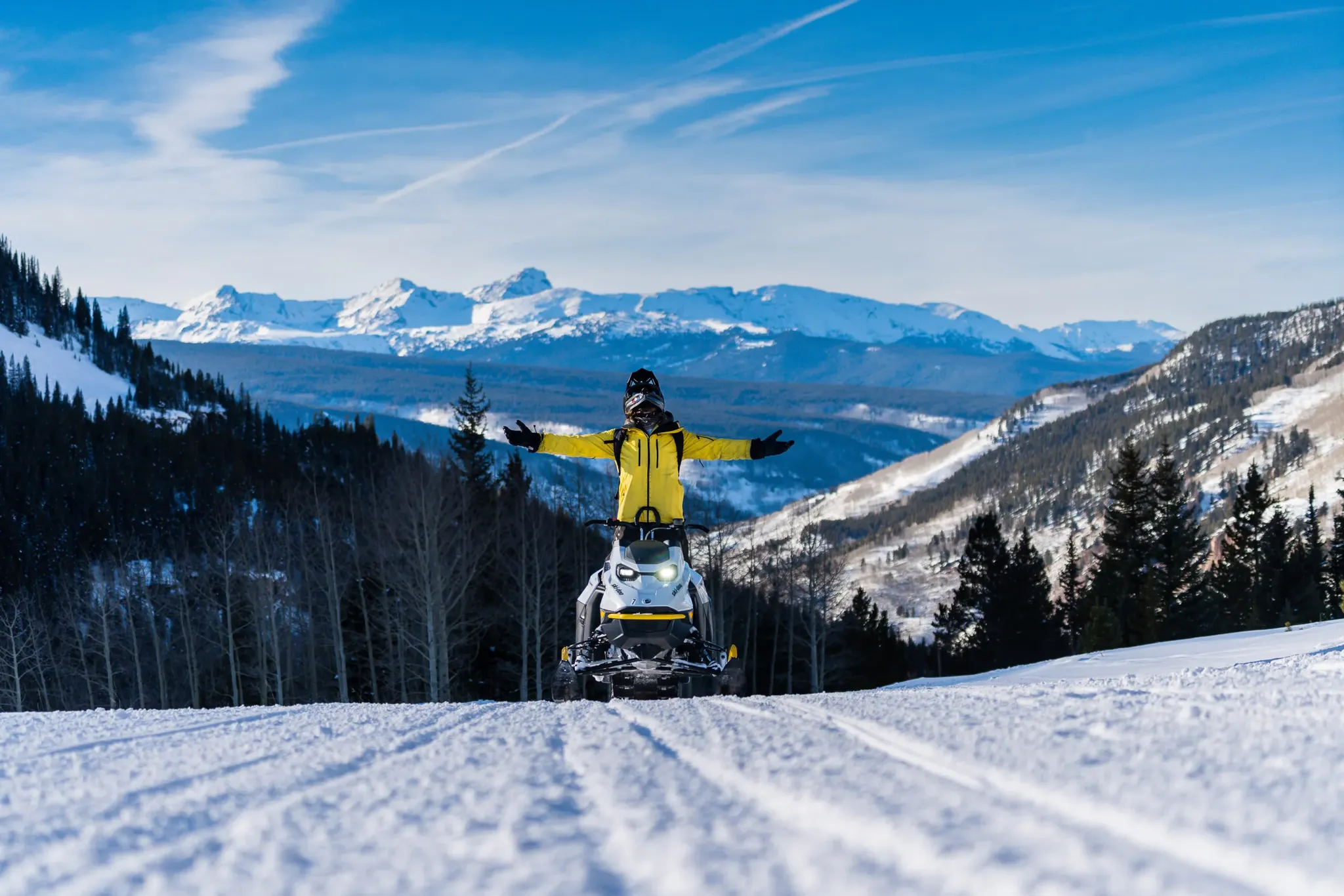 rider standing on snowmobile with arms flung open in front of sweeping mountains views. 