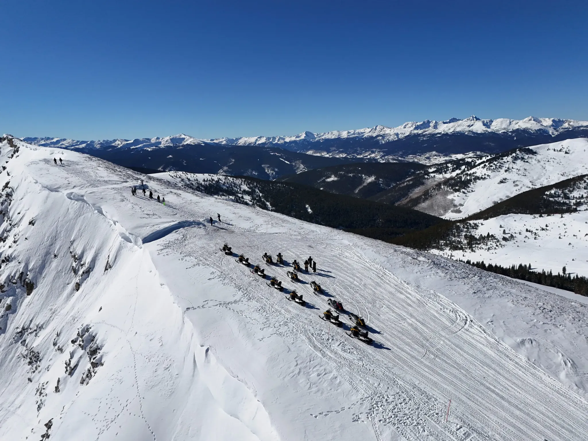 group of snowmobiles parked on snowy mountainside while riders walk to see sweeping mountain views.