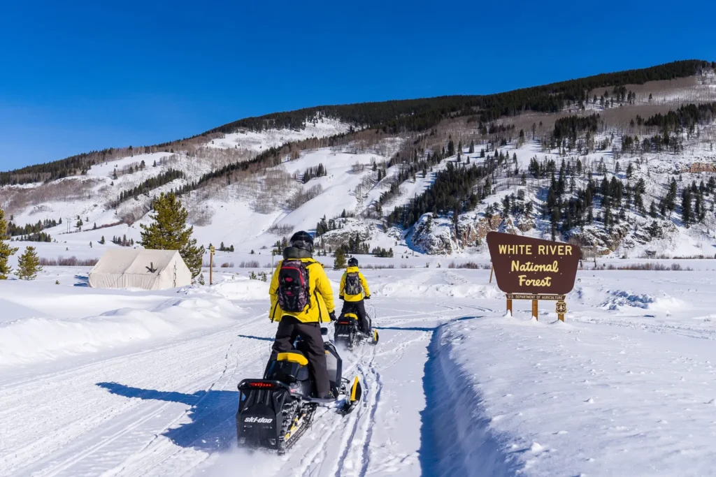 snowmobile riders approaching mountain next to trail sign that reads white river national forest.