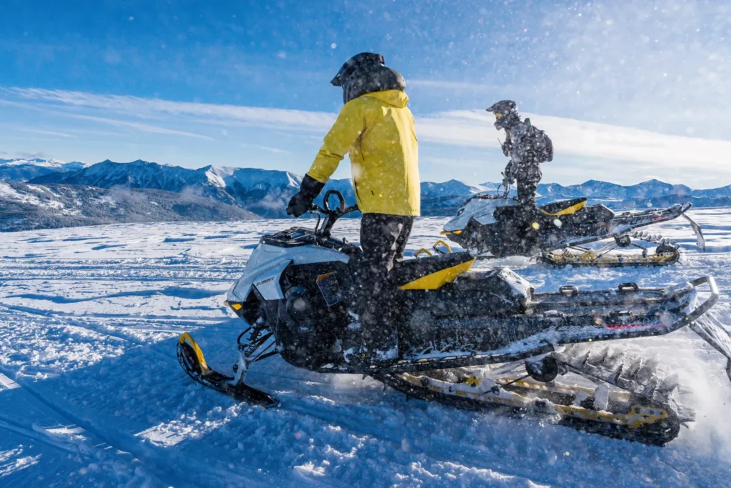 two snowmobile riders standing up on their snowmobiles on flat snowy landscape.
